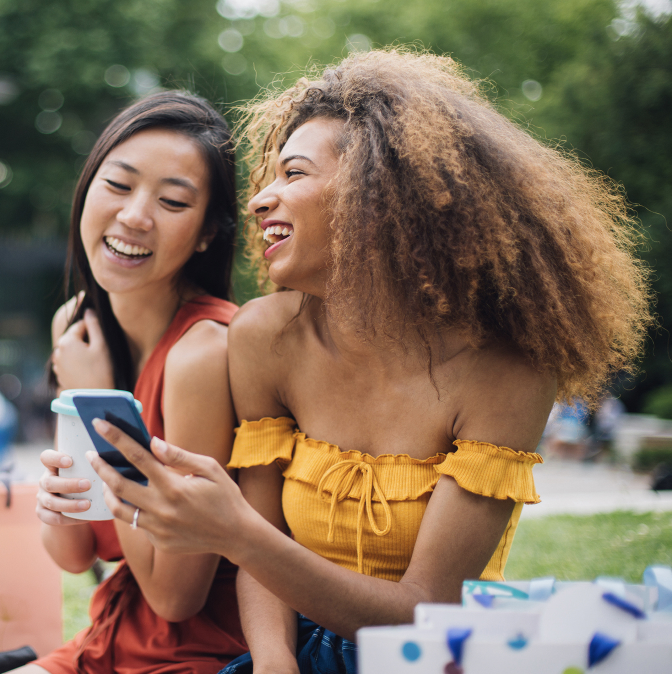 Two teenage Patelco members sitting outside and laughing at what they're seeing on a mobile phone